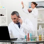 A pair of scientists conducting an experiment. The man sits over a computer whilst a woman scientist is viewing a pipette reading. Coloured test-tubes sit on the table.