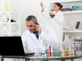 A pair of scientists conducting an experiment. The man sits over a computer whilst a woman scientist is viewing a pipette reading. Coloured test-tubes sit on the table.