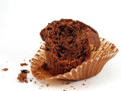 A half eaten chocolate muffin in a case, on a white background.
