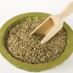 An olive green bowl of hemp seeds with a scoop inserted into the middle. White background.