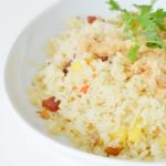 A mixed vegetable rice dish with a sprg of green herb on top, in a white bowl on a white background.