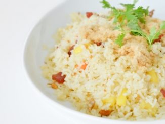A mixed vegetable rice dish with a sprg of green herb on top, in a white bowl on a white background.