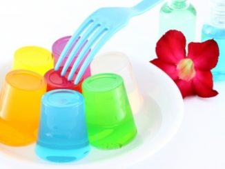 Multicoloured jellies made with meat or fish gelatin, a blue fork, an Allamanda flower and some bottles of coloured liquid. All on a white background.