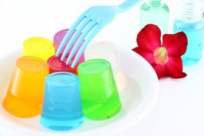 Multicoloured jellies made with meat or fish gelatin, a blue fork, an Allamanda flower and some bottles of coloured liquid. All on a white background.