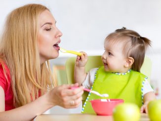 Little girl and her mother with baby food feeding each other, sitting at table in nursery