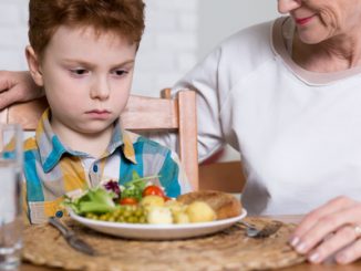 Close-up of a little boy looking malevolently at a dinner plate full of healthy food with his mother looking kindly on.