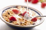 A bowl of cereal containing red pieces of fruit, a spoon and some milk being poured on the product.