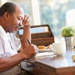 Senior man writing memoirs in book sitting at desk.