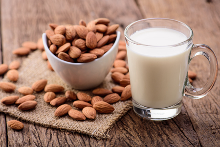 Almonds in a white bowl, almond milk in a glass with handle on a wooden table.