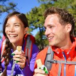 Outdoors couple eating muesli bar hiking. Happy people enjoying granola cereal bars living healthy active lifestyle in mountain nature. Woman and man hiker sitting laughing during hike.