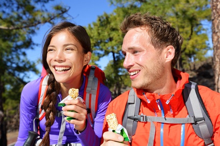 Outdoors couple eating muesli bar hiking. Happy people enjoying granola cereal bars living healthy active lifestyle in mountain nature. Woman and man hiker sitting laughing during hike.