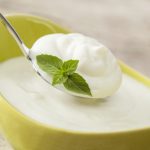 Yogurt in a yellow bowl with some on a silver spoon. Often produced using a Lactobacillus culture.