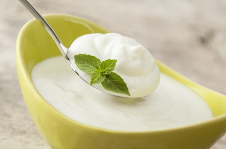 Yogurt in a yellow bowl with some on a silver spoon. Often produced using a Lactobacillus culture.