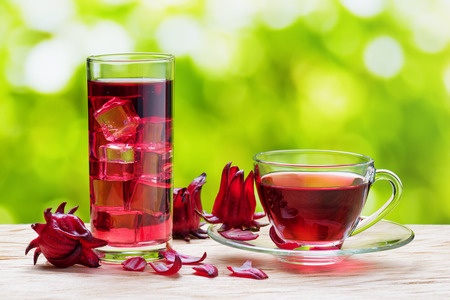 Cup of hot hibiscus tea (karkade, red sorrel, agua de flor de jamaica) and the same cold drink with ice cubes in glass on nature background. magenta-color calyces (sepals) of roselle flowers on table.