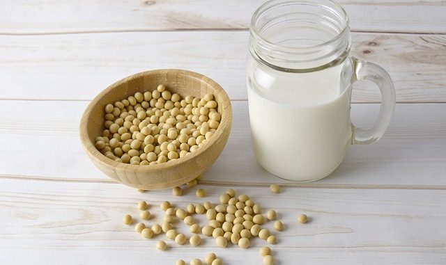 Soy milk in a jug, soy beans in a brown bowl and all on a white table.