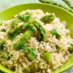 A risotto with asparagus in a pale green bowl on a green cloth with asparagus.