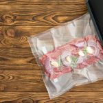 Vacuum sealer. Raw red meat with union and garlic inside plastic wrap next to black kitchenware machine over wood grain background.