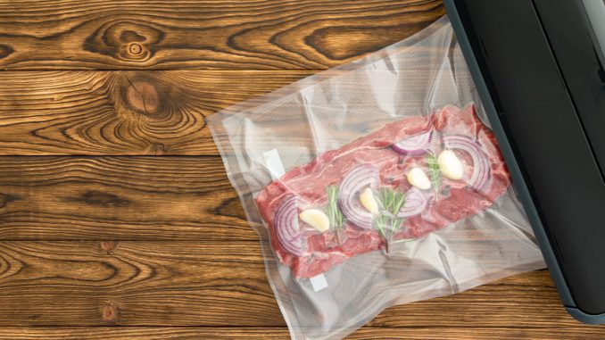 Vacuum sealer. Raw red meat with union and garlic inside plastic wrap next to black kitchenware machine over wood grain background.
