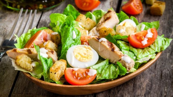 Caesar salad with croutons, quail eggs, cherry tomatoes and grilled chicken in wooden plate on dark rustic table