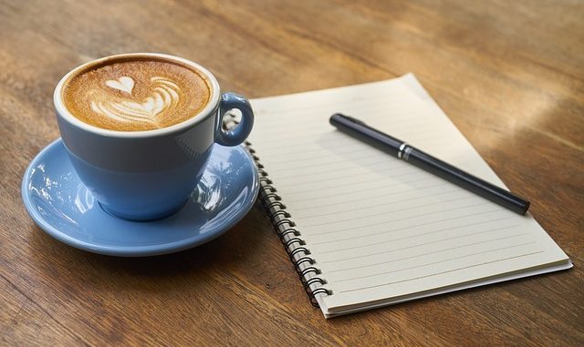 Coffee in a blue cup with saucer, a notebook and pen.