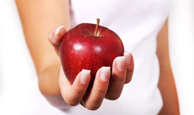 The Rosemary Conley diet may not just rely on an apple in the hand. The person is wearing a white t-shirt. The woman has a very nice manicured set of fingers.
