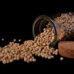 White pepper as corns coming from a glass pot on its side against a black background.