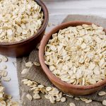 Oat flakes in ceramic bowl and wooden spoon and spikelets on white vintage wooden background, selective focus copy space, top view