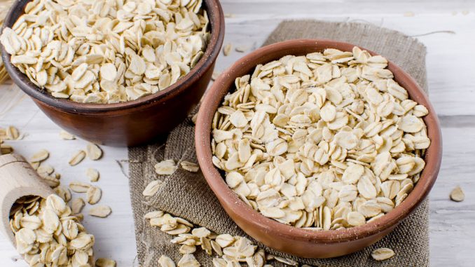 Oat flakes in ceramic bowl and wooden spoon and spikelets on white vintage wooden background, selective focus copy space, top view