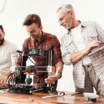 Three men are working on preparing a 3d printer for printing. Two men set up a 3d printer, an elderly man holds a laptop in his hands and watches the process. (3D Printing)