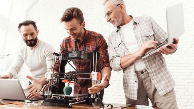 Three men are working on preparing a 3d printer for printing. Two men set up a 3d printer, an elderly man holds a laptop in his hands and watches the process. (3D Printing)