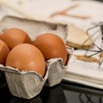 Eggs in a carton on a table next to a whisk.
