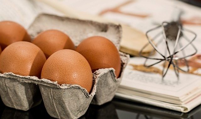 Eggs in a carton on a table next to a whisk.