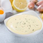 Homemade basic french sauce bearnaise in a white bowl with ingredients, butter, shallot, lemon, eggs, on a light blue stone background, close-up
