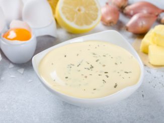 Homemade basic french sauce bearnaise in a white bowl with ingredients, butter, shallot, lemon, eggs, on a light blue stone background, close-up