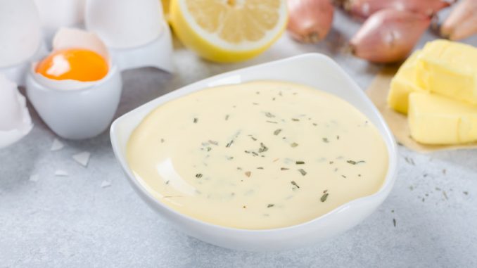 Homemade basic french sauce bearnaise in a white bowl with ingredients, butter, shallot, lemon, eggs, on a light blue stone background, close-up