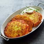 High Angle Still Life of Golden Crisp Fried Potato Rosti Pancakes Served in Dish with Creamy Dill Sauce on Textured Grey Counter Surface.