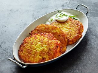 High Angle Still Life of Golden Crisp Fried Potato Rosti Pancakes Served in Dish with Creamy Dill Sauce on Textured Grey Counter Surface.