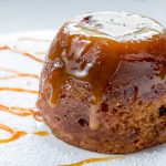 Sticky toffee pudding close up on a white sugar dusted plate drizzled with caramel sauce.