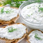 Homemade greek tzatziki sauce in a glass bowl with ingredients and sliced bread on a dark black stone background. Cucumber, lemon, dill, garlic. Close-up, horizontal image, selective focus on a bowl