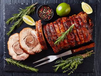 Overhead view of sliced roast pork roulade - Porchetta, delicious pork roast of Italian culinary holiday tradition on a slate tray with rosemary and lime, close-up.