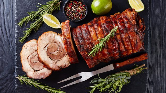 Overhead view of sliced roast pork roulade - Porchetta, delicious pork roast of Italian culinary holiday tradition on a slate tray with rosemary and lime, close-up.