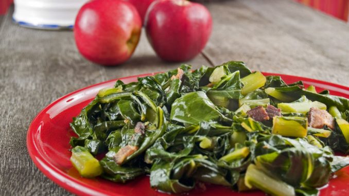 Sautéed greens and bacon on a red plate with apples and a water pitcher in the background.