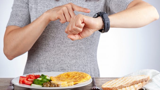 Intermittent fasting concept with a woman sitting hungry in front of food and looking at her watch to make sure she breaks fast on the correct time. A dietary modification for healthy lifestyle. During fasting, gluconeogenesis takes over to generate glucose.