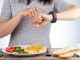 Intermittent fasting concept with a woman sitting hungry in front of food and looking at her watch to make sure she breaks fast on the correct time. A dietary modification for healthy lifestyle. During fasting, gluconeogenesis takes over to generate glucose.