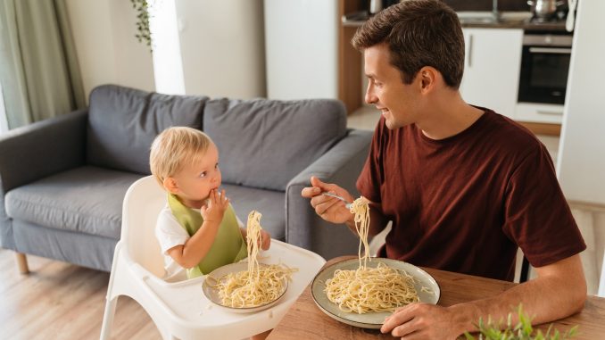 Upper view of dad and baby in high chair eating spaghetti for lunch at kitchen table. Safety assessments are needed for all novel foods.