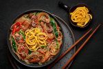 Japchae in black bowl on dark slate table top. Korean cuisine glass chapchae noodles dish with vegetables and meat. Asian traditional food. Authentic meal. Top view. Asian noodles with beef and vegetables in a bowl on a black background
