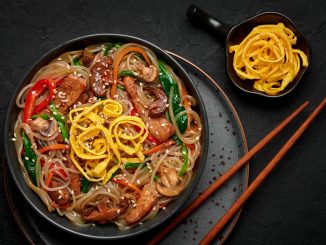 Japchae in black bowl on dark slate table top. Korean cuisine glass chapchae noodles dish with vegetables and meat. Asian traditional food. Authentic meal. Top view. Asian noodles with beef and vegetables in a bowl on a black background