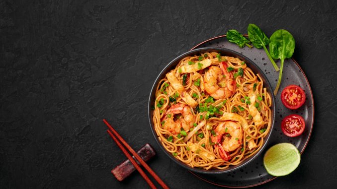 Mie Goreng in black bowl on dark slate table top. Indonesian cuisine prawn noodles and vegetables stir fired dish. Asian food. Asian noodles with shrimps, prawns and vegetables on black background.Top view. Copy space