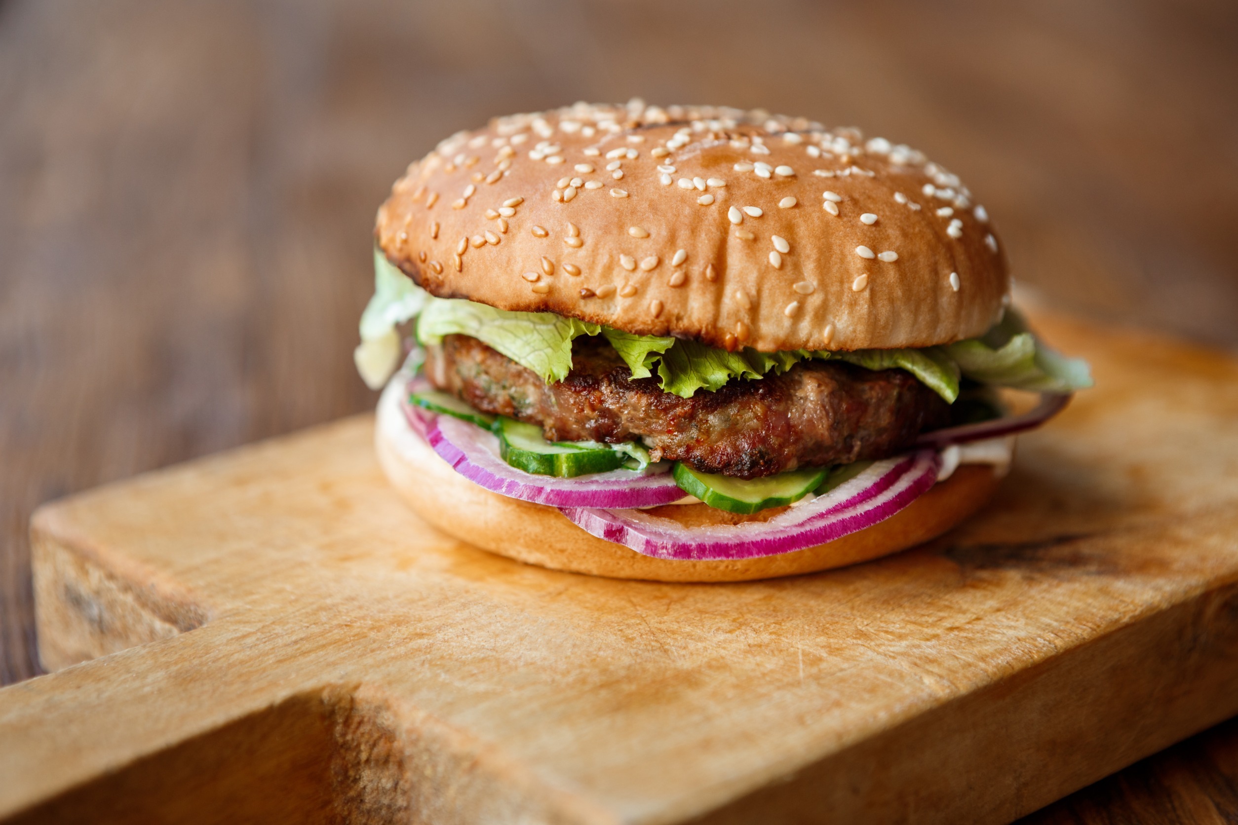 Classic American fast food background. Beefurgers, whole grain bun with grilled on barbecue meat and onions on wooden cutting board. Hamburger with fresh vegetables composition.