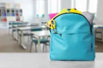 back to school, satchel on table. Light blue backpack with stationery on school desk in classroom. Space for text